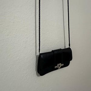 K&ouml;nnte beinhalten: Eine schwarze Leder-Clutch mit einem silbernen herzf&ouml;rmigen Verschluss und einem langen schwarzen geflochtenen Riemen.