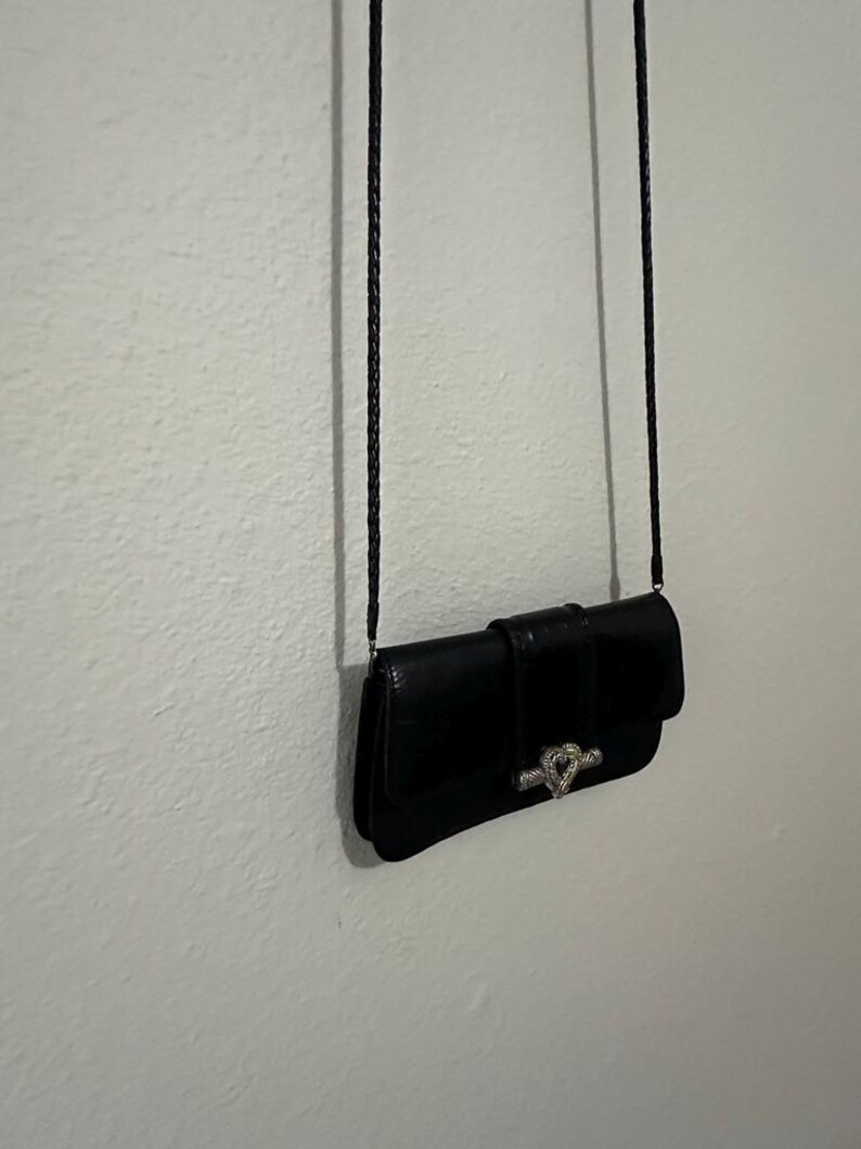 K&ouml;nnte beinhalten: Eine schwarze Leder-Clutch mit einem silbernen herzf&ouml;rmigen Verschluss und einem langen schwarzen geflochtenen Riemen.