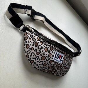 K&ouml;nnte beinhalten: Eine Bauchtasche mit Leopardenmuster, einem schwarzen Rei&szlig;verschluss und einem schwarz-wei&szlig; gestreiften Riemen. Die Tasche hat ein kleines wei&szlig;es Etikett mit der Aufschrift "LOVE BRIGHTON". Die Tasche ist beige, braun und schwarz.