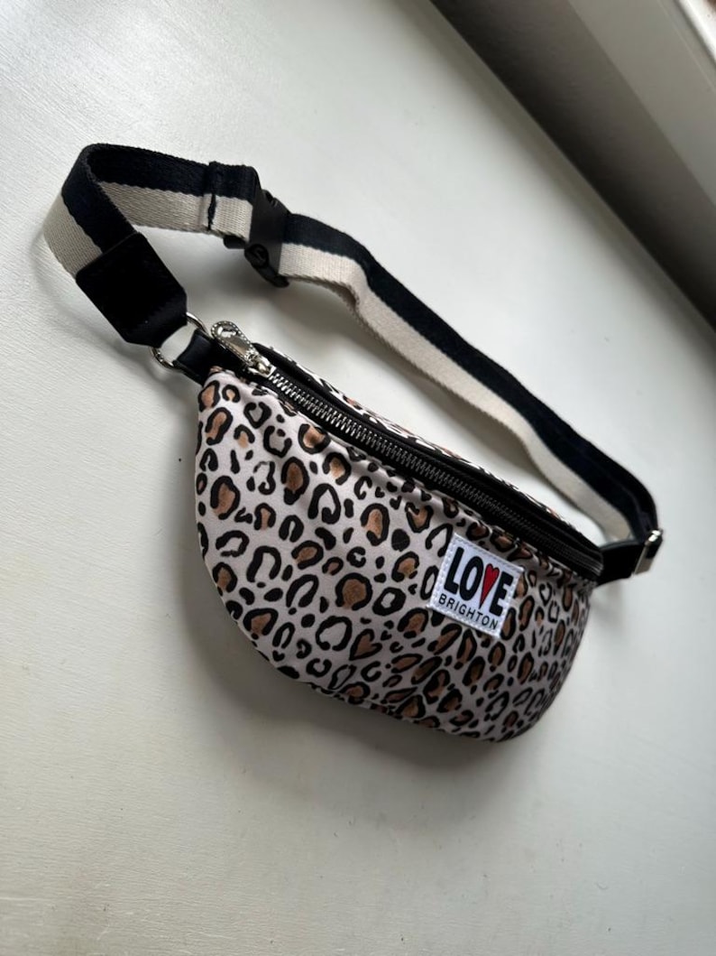 K&ouml;nnte beinhalten: Eine Bauchtasche mit Leopardenmuster, einem schwarzen Rei&szlig;verschluss und einem schwarz-wei&szlig; gestreiften Riemen. Die Tasche hat ein kleines wei&szlig;es Etikett mit der Aufschrift "LOVE BRIGHTON". Die Tasche ist beige, braun und schwarz.
