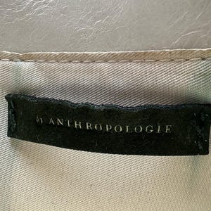 May include: A close-up of a tag that reads "by ANTHROPOLOGIE" in white lettering on a black background. The tag is sewn onto a light-colored fabric with a textured, light gray surface above.