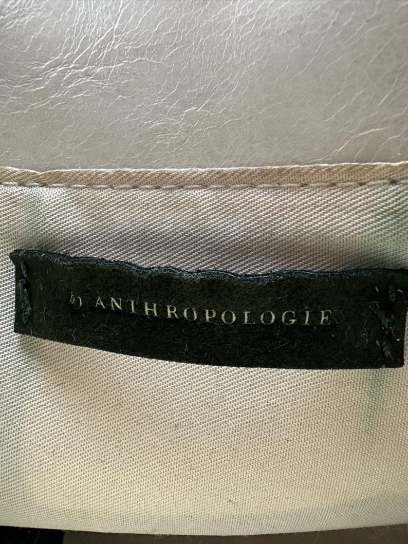 May include: A close-up of a tag that reads "by ANTHROPOLOGIE" in white lettering on a black background. The tag is sewn onto a light-colored fabric with a textured, light gray surface above.