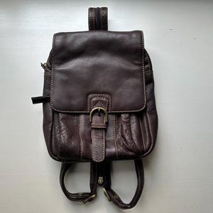 Op de afbeelding: Een donkerbruine leren rugzak met een klep en gespsluiting. De rugzak heeft een klein voorvak en verstelbare bandjes. De stiksels zijn licht van kleur en de hardware is goudkleurig.