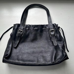 May include: Black leather tote bag with two handles and adjustable straps. The bag features a drawstring closure and silver-tone buckles. The leather has a textured appearance, and the bag is set against a white background.