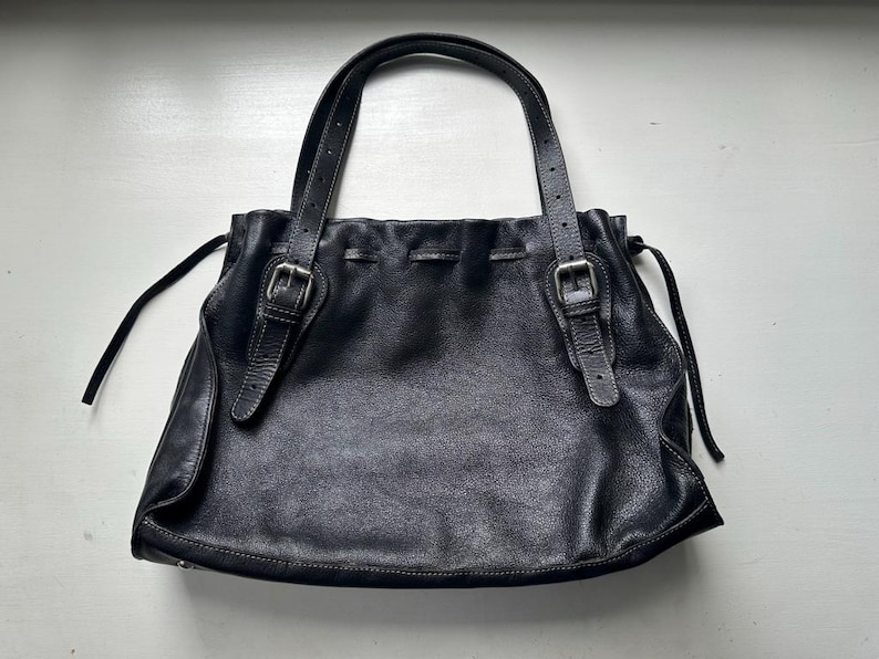 May include: Black leather tote bag with two handles and adjustable straps. The bag features a drawstring closure and silver-tone buckles. The leather has a textured appearance, and the bag is set against a white background.