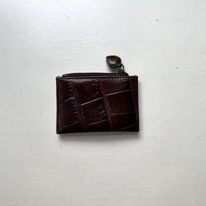 Puede incluir: Una peque&ntilde;a cartera rectangular de cuero marr&oacute;n con un patr&oacute;n texturizado similar al de un cocodrilo. La cartera tiene un cierre de cremallera y una peque&ntilde;a leng&uuml;eta redondeada. Dise&ntilde;ada para guardar tarjetas y efectivo.