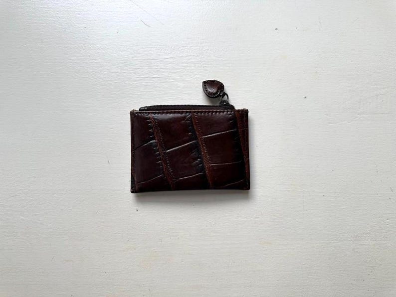 Puede incluir: Una peque&ntilde;a cartera rectangular de cuero marr&oacute;n con un patr&oacute;n texturizado similar al de un cocodrilo. La cartera tiene un cierre de cremallera y una peque&ntilde;a leng&uuml;eta redondeada. Dise&ntilde;ada para guardar tarjetas y efectivo.