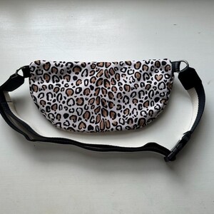 K&ouml;nnte beinhalten: Eine Bauchtasche mit Leopardenmuster und einem verstellbaren schwarzen Riemen und einer Schnalle. Die Tasche hat einen wei&szlig;en Hintergrund mit braunen und schwarzen Flecken. Der Riemen ist schwarz mit einer wei&szlig;en Unterseite.