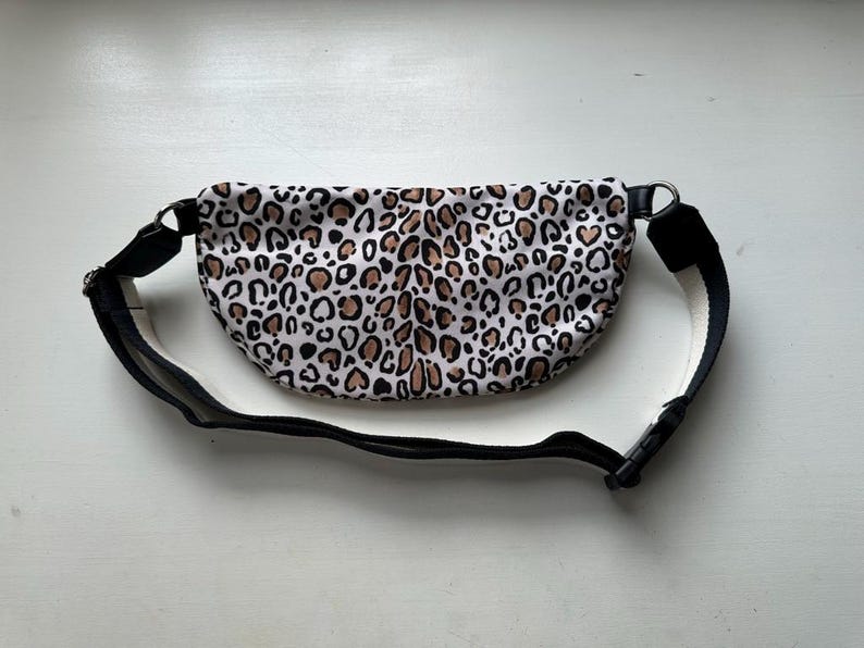 K&ouml;nnte beinhalten: Eine Bauchtasche mit Leopardenmuster und einem verstellbaren schwarzen Riemen und einer Schnalle. Die Tasche hat einen wei&szlig;en Hintergrund mit braunen und schwarzen Flecken. Der Riemen ist schwarz mit einer wei&szlig;en Unterseite.