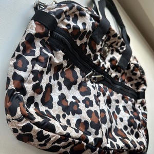 May include: A leopard print travel bag with black straps and zippers. The bag has a white background with brown and black spots.