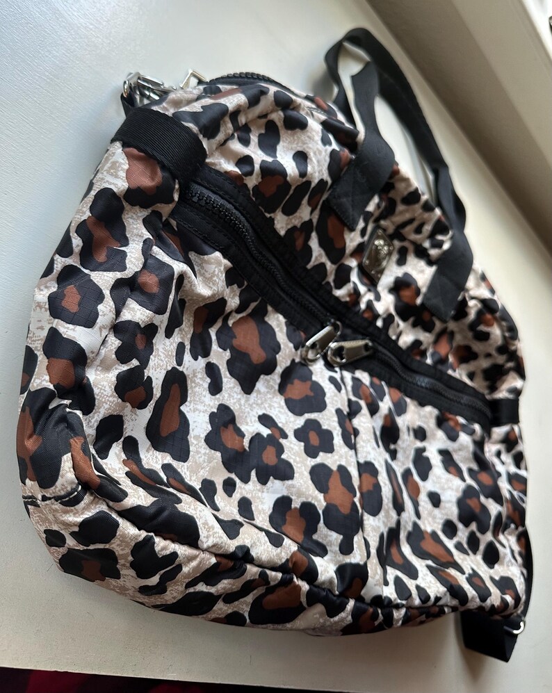 May include: A leopard print travel bag with black straps and zippers. The bag has a white background with brown and black spots.