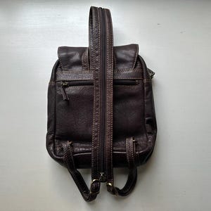 Op de afbeelding: Een donkerbruine leren rugzak met een klepsluiting en een voorvak met ritssluiting. De rugzak heeft verstelbare schouderbanden en een handvat aan de bovenkant. De stiksels zijn lichtbruin en de hardware is messingkleurig.