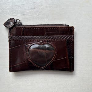 Puede incluir: Una cartera de cuero marr&oacute;n con una ventana en forma de coraz&oacute;n y cierre de cremallera. La cartera tiene un patr&oacute;n texturizado, similar al de un cocodrilo, y varias ranuras para tarjetas. El tirador de la cremallera tambi&eacute;n es marr&oacute;n y en forma de coraz&oacute;n.