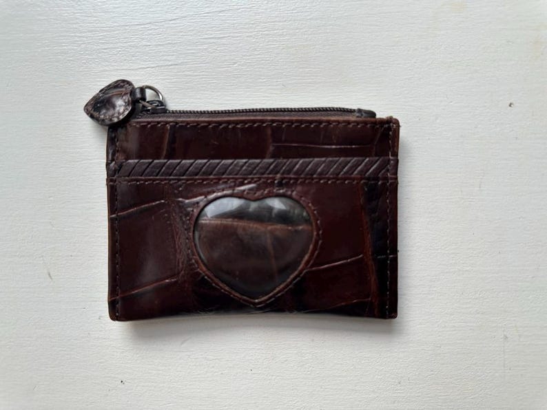 Puede incluir: Una cartera de cuero marr&oacute;n con una ventana en forma de coraz&oacute;n y cierre de cremallera. La cartera tiene un patr&oacute;n texturizado, similar al de un cocodrilo, y varias ranuras para tarjetas. El tirador de la cremallera tambi&eacute;n es marr&oacute;n y en forma de coraz&oacute;n.