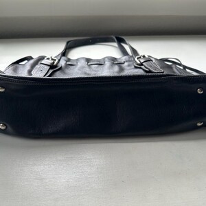 May include: Black leather tote bag with silver-tone hardware. The bag features two top handles, adjustable buckles, and decorative stitching. The bottom of the bag has silver-tone metal feet. The bag is sitting on a white surface.