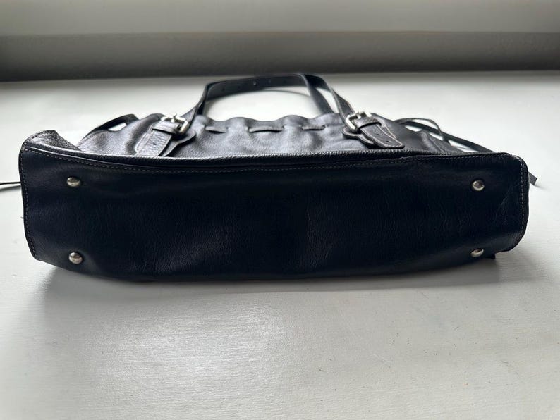 May include: Black leather tote bag with silver-tone hardware. The bag features two top handles, adjustable buckles, and decorative stitching. The bottom of the bag has silver-tone metal feet. The bag is sitting on a white surface.