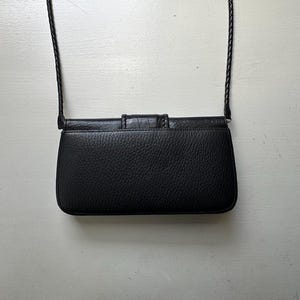 K&ouml;nnte beinhalten: Eine schwarze Leder-Umh&auml;ngetasche mit Klappenverschluss und einem geflochtenen Riemen.