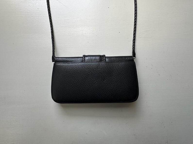 K&ouml;nnte beinhalten: Eine schwarze Leder-Umh&auml;ngetasche mit Klappenverschluss und einem geflochtenen Riemen.