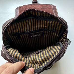 Op de afbeelding: Open zicht op een bruine leren Bianchi crossbodytas. De tas heeft een su&egrave;de flap, een gestreepte binnenvoering en een ritssluiting. Het Bianchi-logo is zichtbaar in de tas.