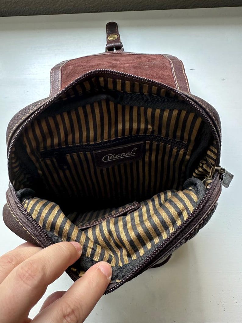 Op de afbeelding: Open zicht op een bruine leren Bianchi crossbodytas. De tas heeft een su&egrave;de flap, een gestreepte binnenvoering en een ritssluiting. Het Bianchi-logo is zichtbaar in de tas.