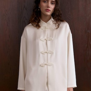 Könnte beinhalten: Cremefarbenes Seiden-Pyjama-Set. Das Oberteil hat einen Stehkragen und dekorative Froschverschlüsse. Das langärmlige Oberteil und die passende Hose schaffen einen entspannten, eleganten Look. Der Stoff hat einen dezenten Glanz.