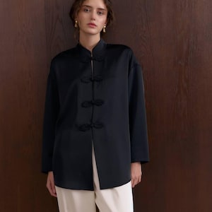 Op de afbeelding: Een zwart overhemd met een mandarijnkraag en decoratieve kikker sluitingen. De top met lange mouwen wordt gecombineerd met een crèmekleurige broek. De outfit wordt gedragen door een persoon voor een donkerbruine houten muur.