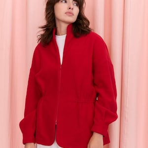 May include: A bright red fleece jacket with a high collar and a full-length zipper. The jacket has long sleeves and a drawstring waist. The model is wearing a white t-shirt and white pants. The background is a pink curtain.