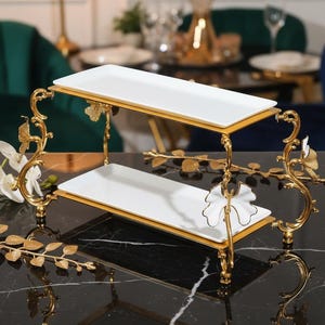 Puede incluir: Una bandeja de servicio de dos niveles con platos rectangulares blancos y detalles metálicos dorados. La bandeja presenta diseños florales y de hojas ornamentados. Los platos están ribeteados con oro. La bandeja está diseñada para servir alimentos.