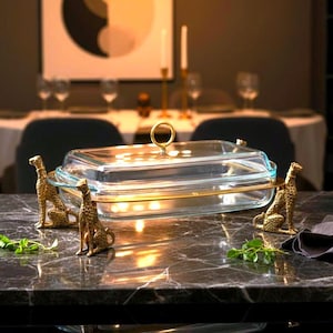 May include: A rectangular glass serving dish with a gold-coloured metal handle and trim. The dish is supported by four gold-coloured cheetah figurines. The dish is on a dark marble surface.