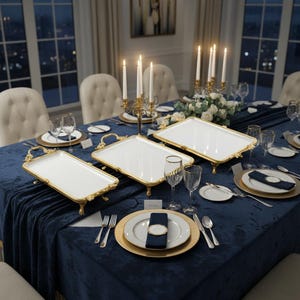 Może przedstawiać: Elegancki stół jadalny na formalne wydarzenie. Stół pokryty jest ciemnoniebieskim aksamitnym obrusem i nakryty białymi talerzami, złotymi podkładkami i złotymi tacami do serwowania. Wysokie białe świece w złotych świecznikach i kieliszki do wina.