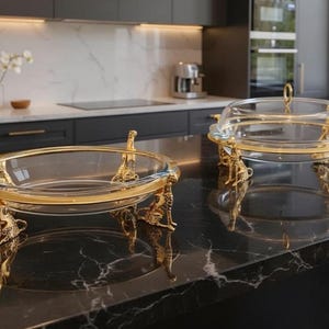 Op de afbeelding: Twee glazen serveerschalen met goudkleurige metalen standaards. Eén schaal heeft een doorzichtig glazen deksel met een gouden handvat. De standaards hebben sierlijke, decoratieve poten. De schalen staan op een zwart aanrechtblad in een keuken.