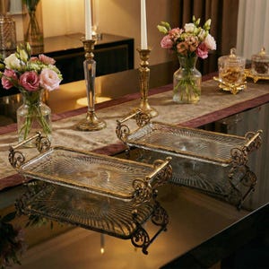 Peut inclure: Deux plateaux rectangulaires en verre avec des cadres et des poignées en métal doré ornementé. Les plateaux présentent un motif en forme d'éventail. La scène comprend des vases de fleurs, des bougeoirs et des bocaux décoratifs, suggérant un thème de décoration intérieure.