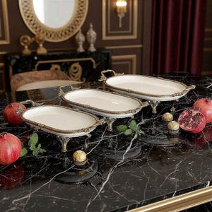 Puede incluir: Tres bandejas ovaladas con interiores blancos y bordes y asas doradas ornamentadas. Las bandejas están sobre patas decorativas doradas. Sobre una superficie de mármol negro, con granadas y bombones.