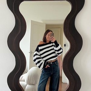 Può includere: Uno specchio marrone scuro con bordi ondulati riflette una persona che indossa un maglione a righe bianche e nere e jeans blu scuro. Lo specchio ha un design unico e stratificato. La persona tiene in mano un telefono.