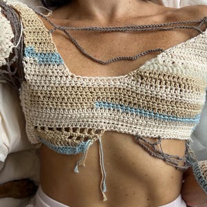 May include: Crocheted top in cream, tan, and light blue. The top features long sleeves with a net-like design and a unique open-front style. The handmade garment is a bohemian style, perfect for summer.