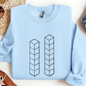 May include: A light blue sweatshirt featuring two vertical columns of stacked cube shapes in black. The sweatshirt has a crew neck and long sleeves. The design is simple and geometric.
