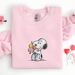 Puede incluir: Una sudadera rosa claro con una imagen bordada de Snoopy sosteniendo un ramo de flores. El personaje de dibujos animados es en blanco y negro, con una oreja negra y una mancha negra en la espalda. Las flores son amarillas, naranjas y rosas.