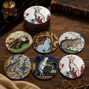 Könnte beinhalten: Ein Set aus runden Untersetzern mit verschiedenen Katzenillustrationen. Die Motive zeigen Katzen in mittelalterlich anmutenden Szenen, einige mit Kronen, andere inmitten von Blumenmustern und eine Katze auf einem blauen Sternenhintergrund.