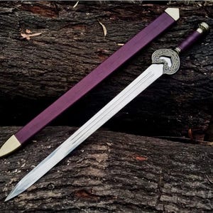 May include: A silver sword with a decorative handle and a purple sheath with gold accents. The sword is lying on a dark brown, textured surface, showcasing its sharp blade and ornate design. The sword's handle is a deep purple color.