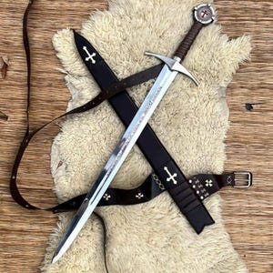 Pode incluir: Uma espada medieval com uma lâmina prateada e um cabo de couro castanho, numa bainha preta. A bainha é adornada com cruzes brancas e um cinto de couro castanho. A guarda da espada apresenta um emblema circular.
