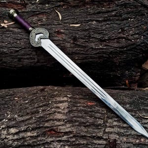 May include: A silver sword with a decorative hilt and a purple handle rests on a dark brown, textured surface. The sword features a long, double-edged blade with three parallel grooves. The hilt has an ornate, circular design.