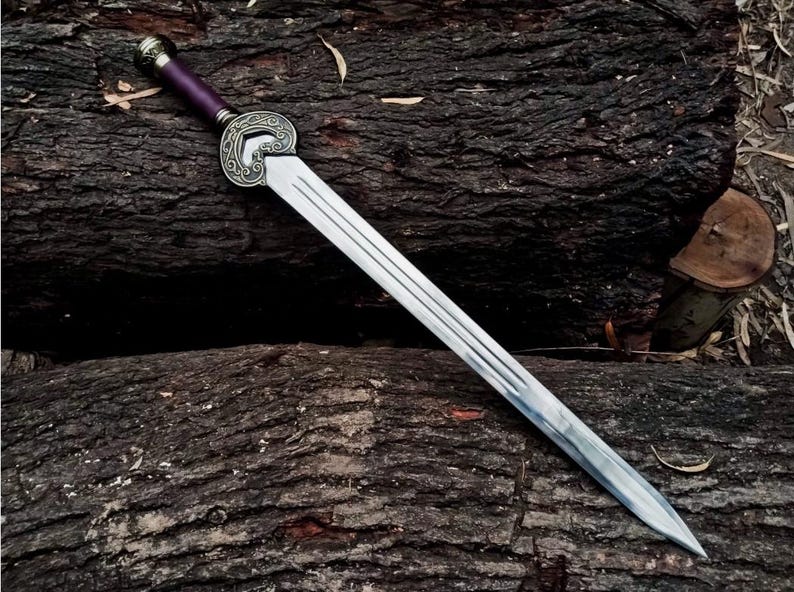 May include: A silver sword with a decorative hilt and a purple handle rests on a dark brown, textured surface. The sword features a long, double-edged blade with three parallel grooves. The hilt has an ornate, circular design.