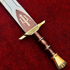 Op de afbeelding: Een gedetailleerd beeld van een zwaard met een zilveren lemmet en een bruin en gouden handvat. De pareerstang toont een drietandsymbool en het Griekse woord "ANAKAYΣMOΣ". Het zwaard ligt op een rode ondergrond.