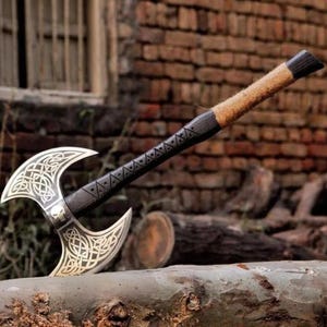 Könnte beinhalten: Eine zweischneidige Axt mit silberfarbenem Kopf und dunklem Holzgriff. Der Axtkopf weist komplizierte keltische Knotenmuster und ein Totenkopfdetail auf. Der Griff ist mit beigefarbenem Garn umwickelt.