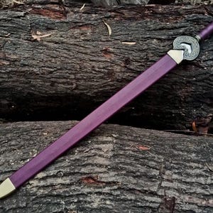 May include: A purple sword with a gold-colored tip and a decorative silver hilt. The sword is resting on a bed of dark brown logs, creating a rustic aesthetic. The sword's handle is also purple.