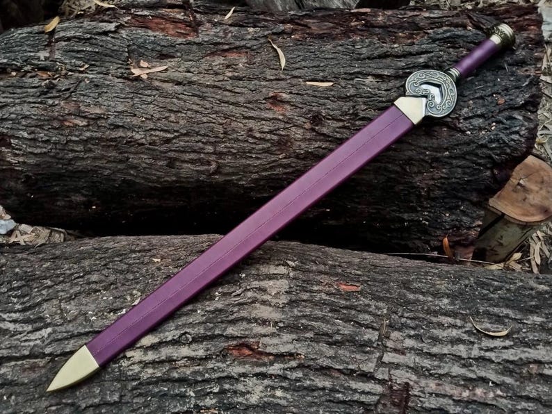 May include: A purple sword with a gold-colored tip and a decorative silver hilt. The sword is resting on a bed of dark brown logs, creating a rustic aesthetic. The sword's handle is also purple.