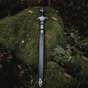May include: A detailed image of a Viking sword with a black scabbard and ornate silver-colored hilt and crossguard. The sword rests on a moss-covered rock, with foliage in the background. The sword's design features intricate patterns.