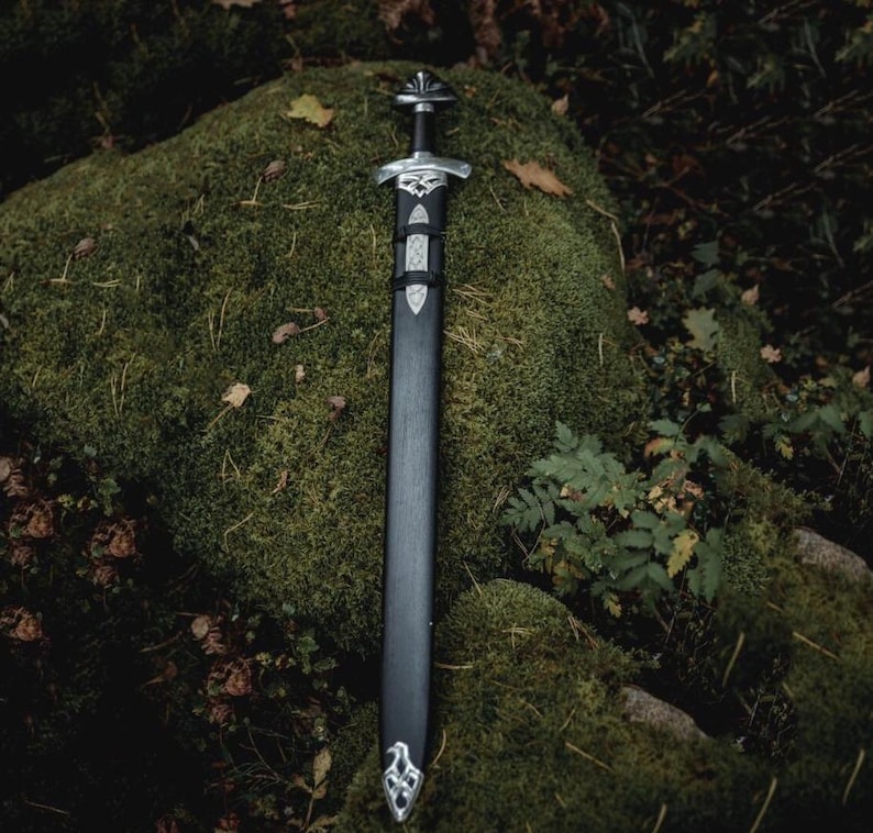 May include: A detailed image of a Viking sword with a black scabbard and ornate silver-colored hilt and crossguard. The sword rests on a moss-covered rock, with foliage in the background. The sword's design features intricate patterns.