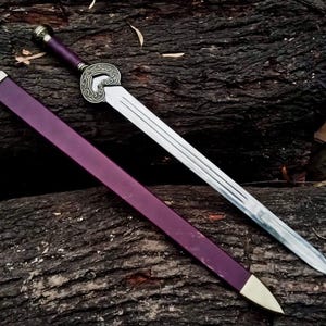 May include: A decorative sword with a silver blade and a purple handle and sheath. The handle features an ornate silver design. The sheath has a gold-colored tip. The sword is resting on a dark brown wooden surface.