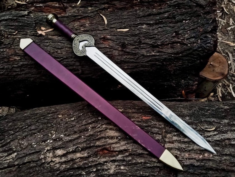 May include: A decorative sword with a silver blade and a purple handle and sheath. The handle features an ornate silver design. The sheath has a gold-colored tip. The sword is resting on a dark brown wooden surface.